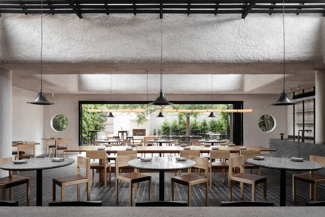 The dining room at Arkhé in Norwood, timber tables beneath textured plaster with the leafy courtyard beyond