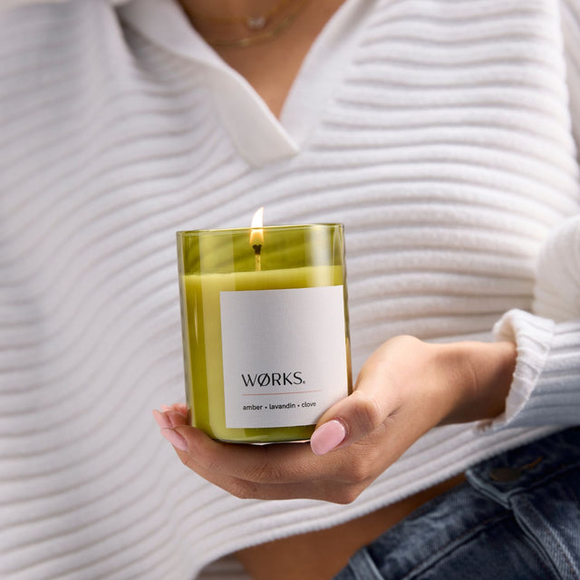 Model relaxing beside WØRKS Amber Candle in green glass — warm and resinous with amber, lavandin, and clove bud.