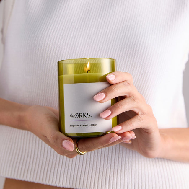 Model lighting WØRKS Bergamot Candle in deep green glass — a citrus-forward blend of bergamot, petitgrain, and cedar.