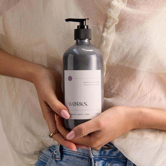 Model applying WØRKS Bergamot Hand Cream from a 500ml glass bottle — richly hydrating with bergamot, petitgrain, and cedar.