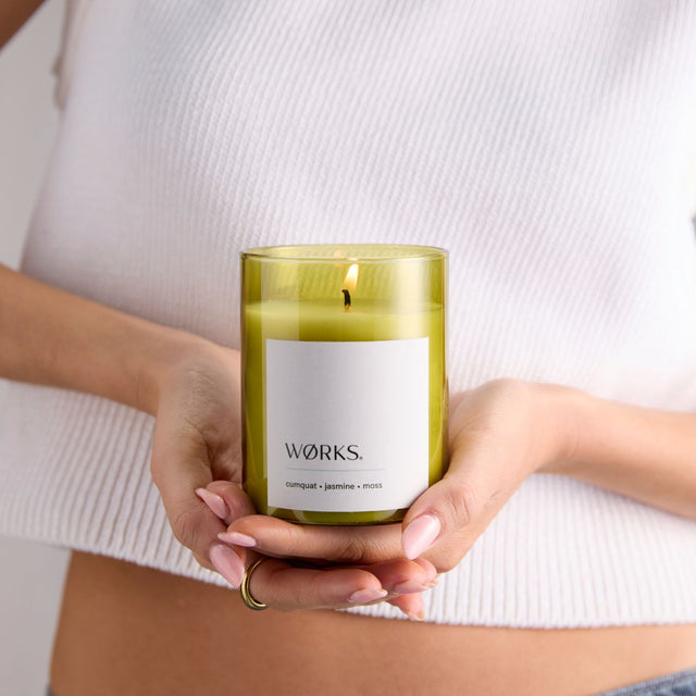 Model holding WØRKS Cumquat Candle in green glass vessel — a fresh, floral blend of cumquat, jasmine, and moss.