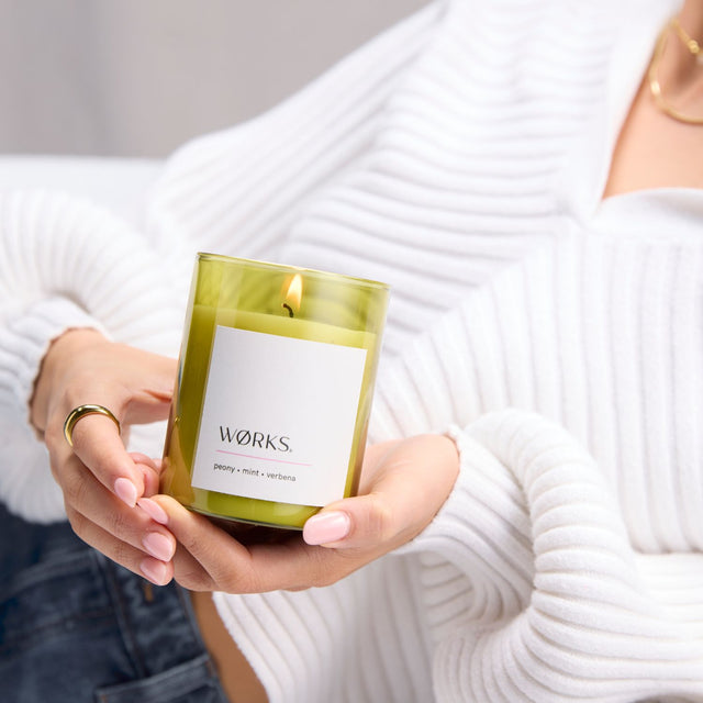 Model placing WØRKS Peony Candle on table in green glass — a soft, refreshing blend of peony, mint, and verbena.