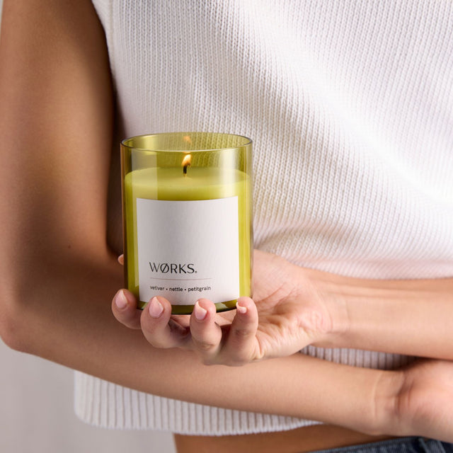Model enjoying the scent of WØRKS Vetiver Candle in green glass — grounding notes of vetiver, nettle, and petitgrain.