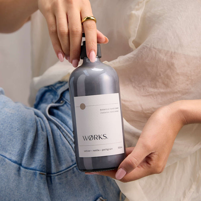 Model using WØRKS Vetiver Hand Cream in 500ml glass bottle — deep moisture with vetiver, nettle, and petitgrain.