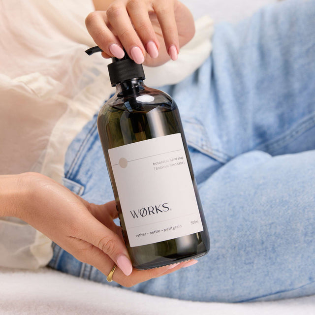 Model washing hands with WØRKS Vetiver Hand Soap in 500ml smoky glass bottle — grounding notes of vetiver, nettle, and petitgrain.