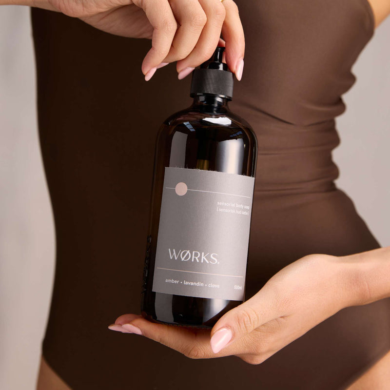 Model using WØRKS Amber Body Soap in 500ml smoky glass bottle, warm notes of amber, lavandin, and clove bud.
